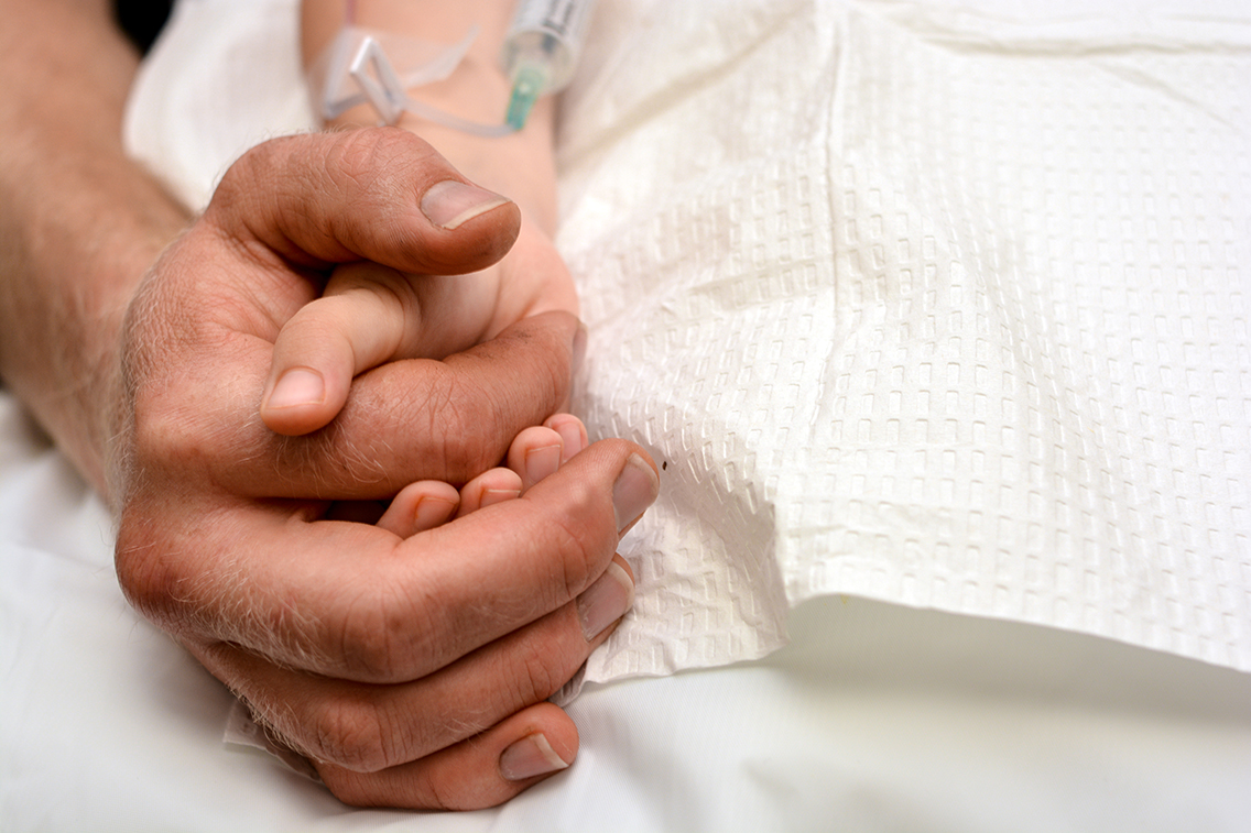 Close-up of a father holding his son's hand during a medical treatment.